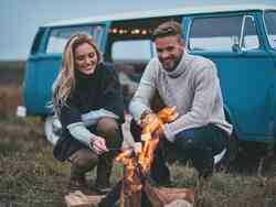 Ein Mann und eine Frau machen ein Lagerfeuer. Dahinter steht ein blauer VW Bus.